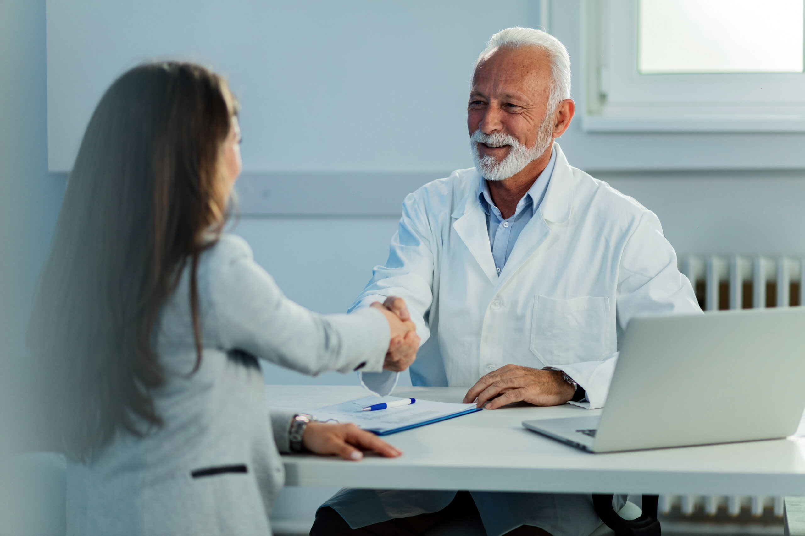 Happy mature doctor and his female patient shaking hands after medical appointment at clinic.